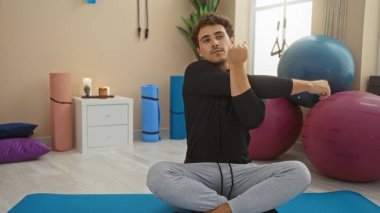 Yoga minderleri ve egzersiz toplarıyla modern bir spor salonunda esneyen genç bir adam, rahat bir egzersiz atmosferi sergiliyor..