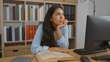 Genç bir kadın düşünceli bir şekilde çalışma odasındaki bir masada oturuyor. Yanında bir bilgisayar ve açık kitaplarla, kütüphane raflarıyla çevrili..