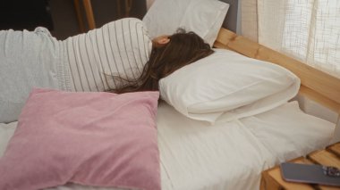 Beyaz yastıklar, pembe yastıklar ve yatak odasında ahşap bir komodin bulunan esmer at kuyruklu bir evde uyuyan bir kadın.