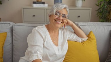 Sarı yastıklı, rahat bir oturma odasında gülümseyen gri saçlı kadın, ev ortamında orta yaşlı, güzel, olgun bir insanı sergiliyor..