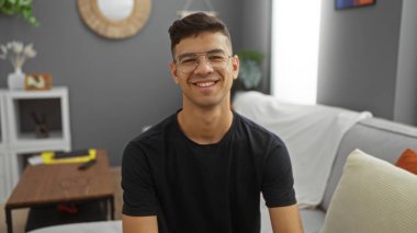 Kapalı bir oturma odasında gülümseyen genç bir adam çekici ve dostane bir tavır sergiliyor..