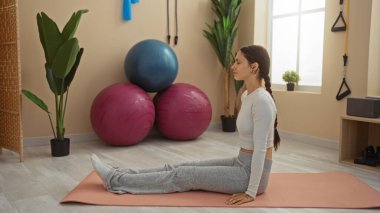 Spor salonunda yoga yapan genç bir kadın. Etrafı spor topları ve parlak bir odadaki kapalı hava bitkileriyle çevrili.