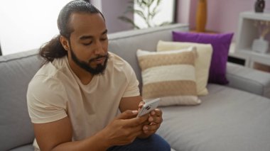 Sakallı genç adam, modern bir oturma odasında rahat bir atmosferle akıllı telefon kullanıyor ve ev teknolojisini ön plana çıkarıyor..