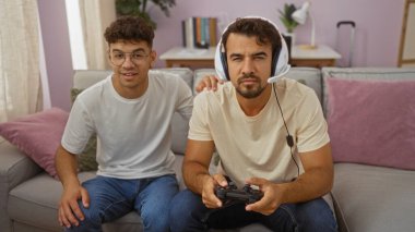 Baba ve oğul birlikte oturma odasında oturuyorlar, ikisi de sıradan kıyafetler giyiyorlar, baba video oyunu oynuyor, kontrol ve kulaklık takıyor, çocuk da izliyor ve gülümsüyor..