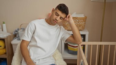 Genç İspanyol bir adam hastane odasında bir beşikte dinleniyor ve doğal ışıklandırmalı klinik bir bebek ortamında düşünceli bir iç atmosfer sergiliyor..