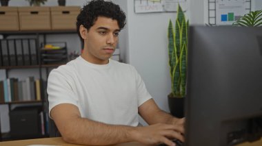 Yakışıklı İspanyol adam, modern bir ofiste bilgisayarına odaklanmış, kitaplıklar ve bitkilerle çevrili, verimli bir iş yeri ortamı örneği..