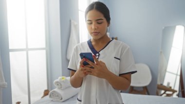 Klinikteki sakin atmosferi olan akıllı telefonu kullanan kadın, üniforma giyiyor, profesyonelliği yansıtıyor ve havlular ve ekipmanlarla sağlık hizmetlerine odaklanıyor..