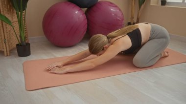 Spor salonundaki minderde yoga yapan genç bir kadın. Arka planda büyük egzersiz topları ve yakınlarda bitkiler var.