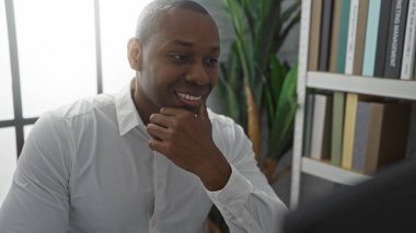 Genç adam parlak bir ofiste gülümsüyor, bir masada oturuyor, kitaplarla dolu bitki ve raflarla çevrili, pozitiflik saçıyor ve profesyonel bir çalışma ortamında odaklanıyor..