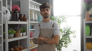 Sakallı ve dövmeli genç İspanyol bir adam bir dekoratif dükkanında vazo, bitki ve ev dekorasyonu malzemeleriyle çapraz duruyor..