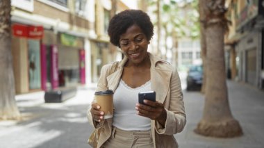 Güzel, orta yaşlı, kıvırcık saçlı, elinde kahve ve akıllı telefonuyla bir şehir caddesinde dururken gülümseyen bir Afrikalı kadın.