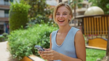 Şehir parkında elinde akıllı telefonuyla gülümseyen bir kadın, şehirde güneşli bir günün tadını çıkartan sıradan kıyafetli sarışın bir kadını gösteriyor.