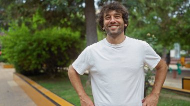Yakışıklı genç adam canlı bir şehir parkında kendine güvenle gülümsüyor güneşli bir günde yeşil ve şehir hayatını gözler önüne seriyor.