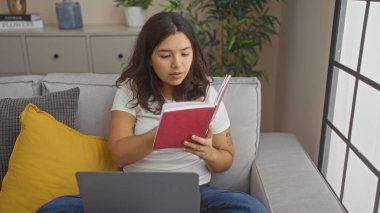 Genç bir İspanyol kadın kucağında dizüstü bilgisayarla oturma odasında otururken kitap okuyor..