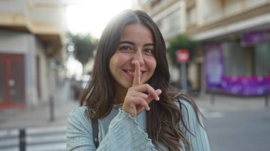 Kentsel sokak ortamında dudakları dudaklarında sessizce gülümseyen kadın, güneş ışığını yansıtan esmer saçlar, şehirde huzurlu ve güzel bir atmosfer yaratıyor..