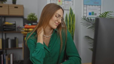 Ofiste boynu ağrıyan, gözlük takan, yeşil bluz giyen, arka planda saksıları olan bir kadın.
