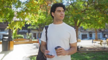 Şehir parkında elinde kahve ve telefonla duran yakışıklı İspanyol bir adam, düşünceli görünüyor..