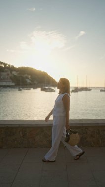 Günbatımında Mallorca, İspanya 'da manzaralı bir rıhtımda yürüyen bir kadın, arka planda yelkenli ve tepe sahil şeridi olan bir çanta tutuyor.