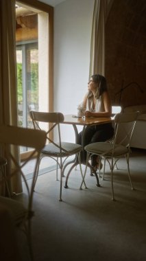 Kapalı bir kafeteryada küçük bir masada oturan gözlüklü genç bir kadın, pencereden dışarı odayı aydınlatan doğal ışıkla bakıyor..