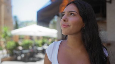 Genç, güzel, esmer bir kadın güneşli bir kasabada şehir sokağında dikiliyor, düşünceli bir ifadeyle başka tarafa bakıyor, latin yüz hatlarını gösteriyor ve sakin bir açık hava anını yakalıyor.