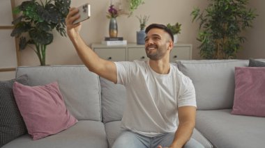 Sakallı, yakışıklı, genç bir İspanyol. Oturma odasında selfie çekiyor. Arka planda yemyeşil bir kanepeye oturuyor..