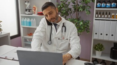 Steteskoplu bir doktor dizüstü bilgisayar ve telefon kullanıyor, kapalı alanda çalışıyor, arka planda organize raflar ve bitkilerle profesyonel tıbbi ortamlar sergiliyor..