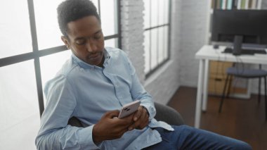 Genç adam ofiste oturuyor, telefon kullanıyor, günlük gömlekler giyiyor, pencerelerden doğal ışık salıyor, modern çalışma alanı içi, rahat atmosfer, odaklanmış ve meşgul..