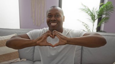 Kapalı alanda gülümseyen Afrikalı genç bir Amerikalı evdeki koltukta oturmuş kalp jestleri yapıyor. Arka planda bitkiler, sıcaklık ve mutluluğu aktarıyor.
