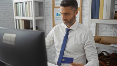 Ofisteki adam, arka planda rafları ve grafikleri olan kredi kartı tutarken, bir işletmeyi veya çalışma düzenini resmeden bilgisayara odaklandı.