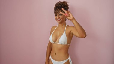 Güzel İspanyol kadın, izole edilmiş pembe arka plan üzerinde beyaz bikinili iki parmağını göstererek zafer işareti yapıyor.