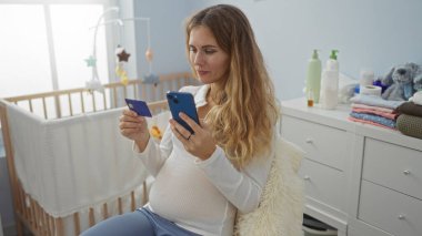 Evin içinde oturan bir kadın, yatak odasının yanında akıllı telefon ve kredi kartı kullanıyor. İnternetten alışveriş ya da bankacılık gibi..