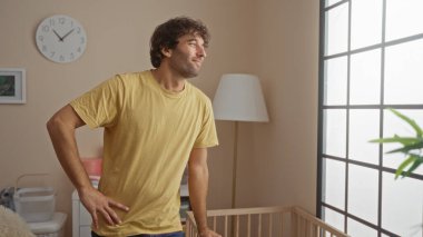 Genç İspanyol bir adam rahat bir yatak odasında, parlak pencereli ve şık dekorlu bir bebek beşiğinin yanında duruyor..