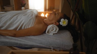 Çiçek saçı aksesuarlı spa yatağında dinlenen bir kadın. Etrafı mum ışığıyla çevrili. Huzur dolu bir sağlık merkezinde.