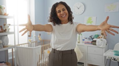 Yatak odasında bebek beşiği ve oyuncaklarıyla sıcacık gülümseyen bir kadın samimi ve davetkar bir atmosfer yaratıyor.