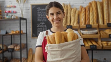 Fırının içinde elinde ekmek tutan genç bir kadın, çeşitli taze pişmiş ürünler sergiliyor, mutlu bir gülümsemeyle..