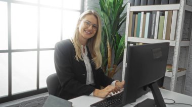 Ofisteki bilgisayarında kitap rafları ve bitkilerle gülümseyen kadın, doğal ışık ile çevrili, profesyonel ve dostane çalışma ortamını vurguluyor.