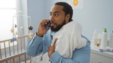 Sakallı bir adam yatak odasında bebeğini tutuyor telefonda konuşuyor, sakin bir ev ortamında beşik ve hafif ışıklandırma atmosferi var..