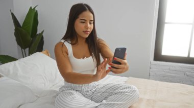 Modern bir yatak odasında yatakta oturan bir kadın, rahat bir iç mekan atmosferi yakalıyor, sakin ve kendinden emin bir ifadeyle Latin güzelliğini vurguluyor..