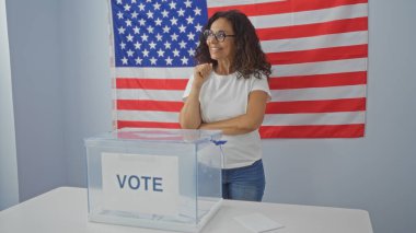 Gülümseyen orta yaşlı İspanyol kadın arka planda Amerikan bayrağı olan bir oy sandığının yanında duruyor..