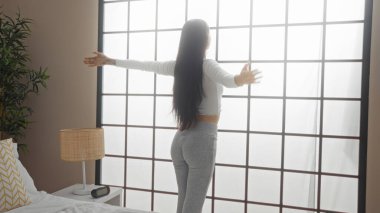 İspanyol kadın güneşli bir yatak odasında büyük bir pencereyle uzanıyor, sabah ışığını kucaklıyor.