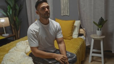 Yatak odasında rahat bir yatakta oturan sakallı genç İspanyol bir adam. Arka planda yastıklar ve bitkiler olan modern bir dekor var..