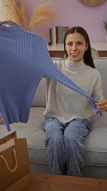 Oturma odasında oturan bir kadın, rahat bir evde, mavi bir süveter ve alışveriş torbalarıyla bir masada oturuyor.