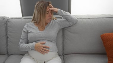 Hamile bir kadın, rahat bir oturma odasında, rahat bir oturma odasında, elleri karnında, rahat giysiler içinde, iç mekan hafifçe aydınlatılmış, bir anlık tefekkür görüntüsü yakalıyor..