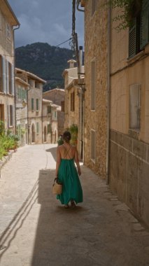 Valldemossa 'nın dar sokaklarında yürüyen kadın Mallorca, İspanya' da, arkasında dağlar olan yeşil bir elbise giyiyor.