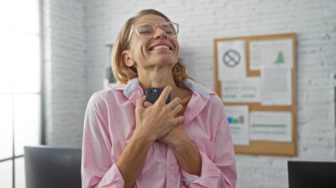 Modern ofis ortamında beyaz tuğladan duvarları olan akıllı telefonu tutan bir kadın mutlu bir şekilde gülümsüyor, iş yerinde mutluluk ve başarı anı yakalıyor..
