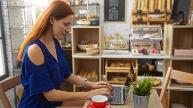 Fırının içinde dizüstü bilgisayarda çalışan genç bir kadın. Etrafında çeşitli hamur işleri sergileniyor.