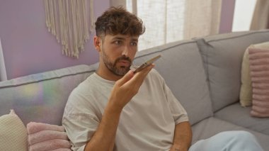 Genç adam evinin oturma odasında akıllı bir telefonla konuşuyor pastel yastıklar ve perdelerin arasından süzülen yumuşak ışıkla sıradan bir iç mekan sergiliyor.