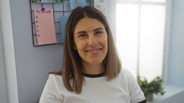 Ofiste gülümseyen ve arka planda yapılacaklar listesi olan genç bir kadın, kapalı alanda pozitif ve profesyonel bir ortam sergiliyor.