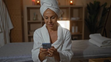 Kaplıcada, kafasında bornoz ve havlu olan bir kadın, arka planda mumlar, raflar ve kapalı alandaki bitkilerle loş ışıklandırılmış bir sağlık odasında akıllı telefon kullanıyor.