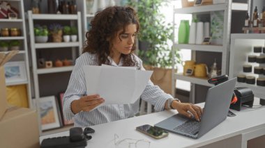 Evrak ve dizüstü bilgisayarla çalışan genç bir kadın güzel dekore edilmiş bir dükkânda ev dekorasyonu malzemeleriyle çalışıyor.